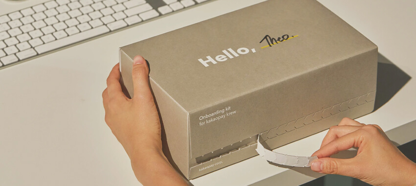 Close-up of hands opening a branded “Hello, Theo” onboarding kit box on a white desk with a computer keyboard nearby.