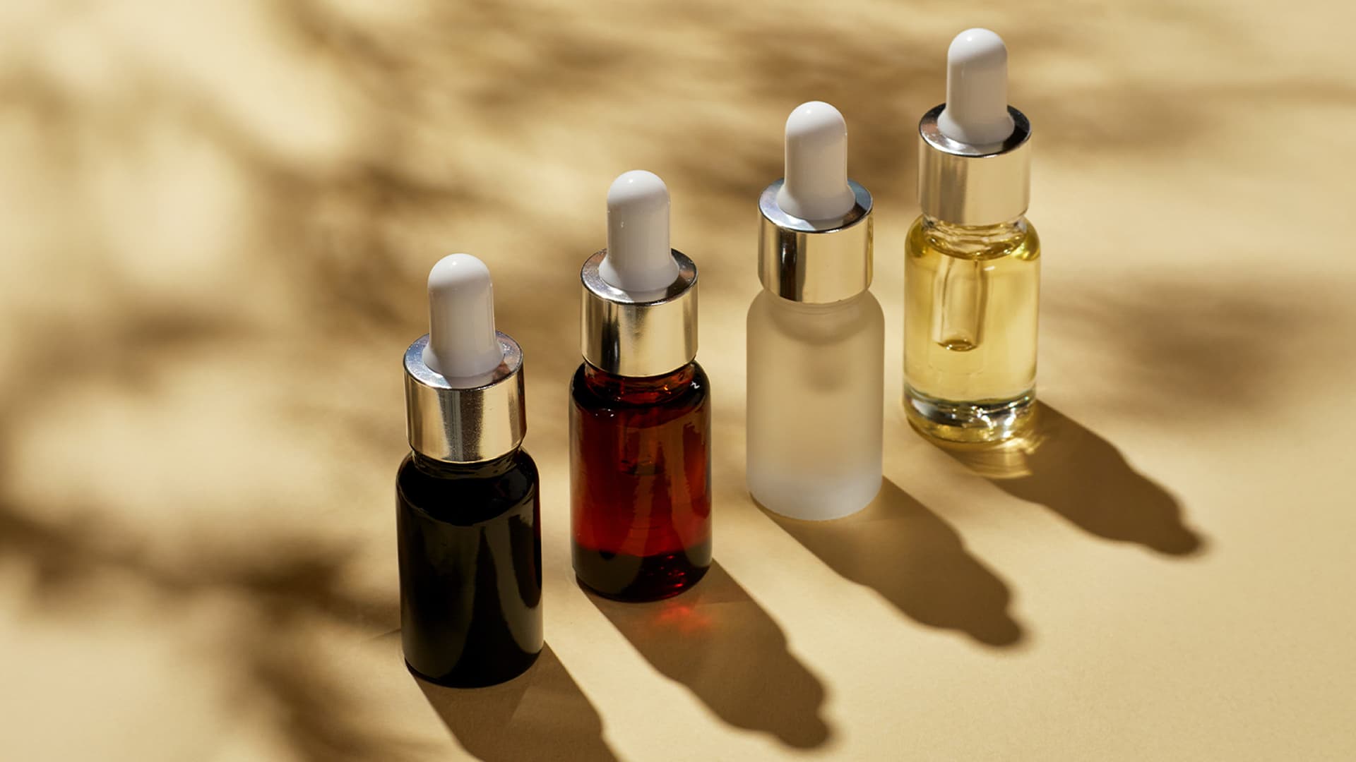 Four glass dropper bottles containing different colored cosmetic liquids are arranged on a beige background with soft shadows.