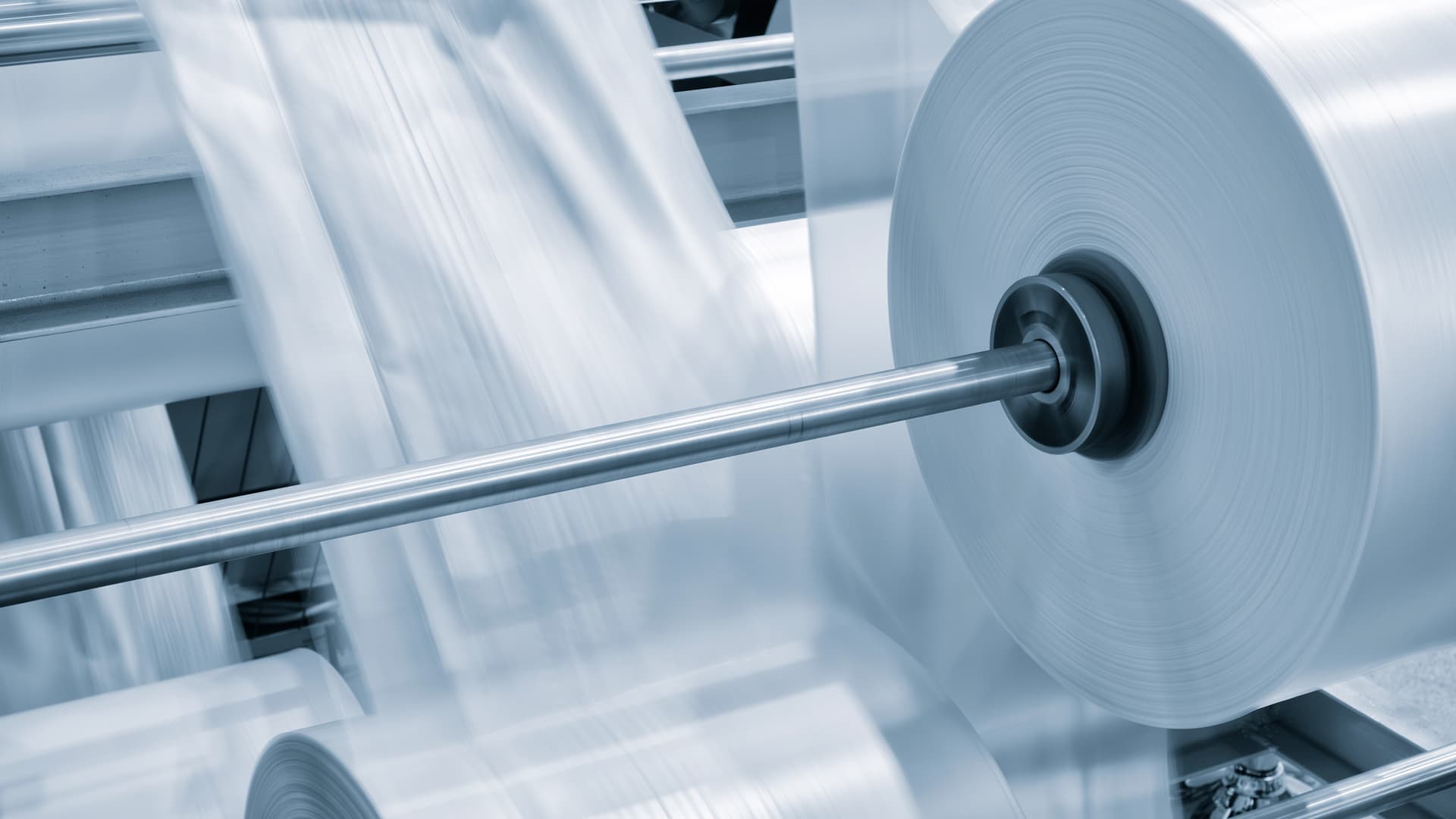 Industrial manufacturing equipment showing a large roll of clear plastic film or material being processed, with motion blur indicating the machinery is in operation during the production process.
