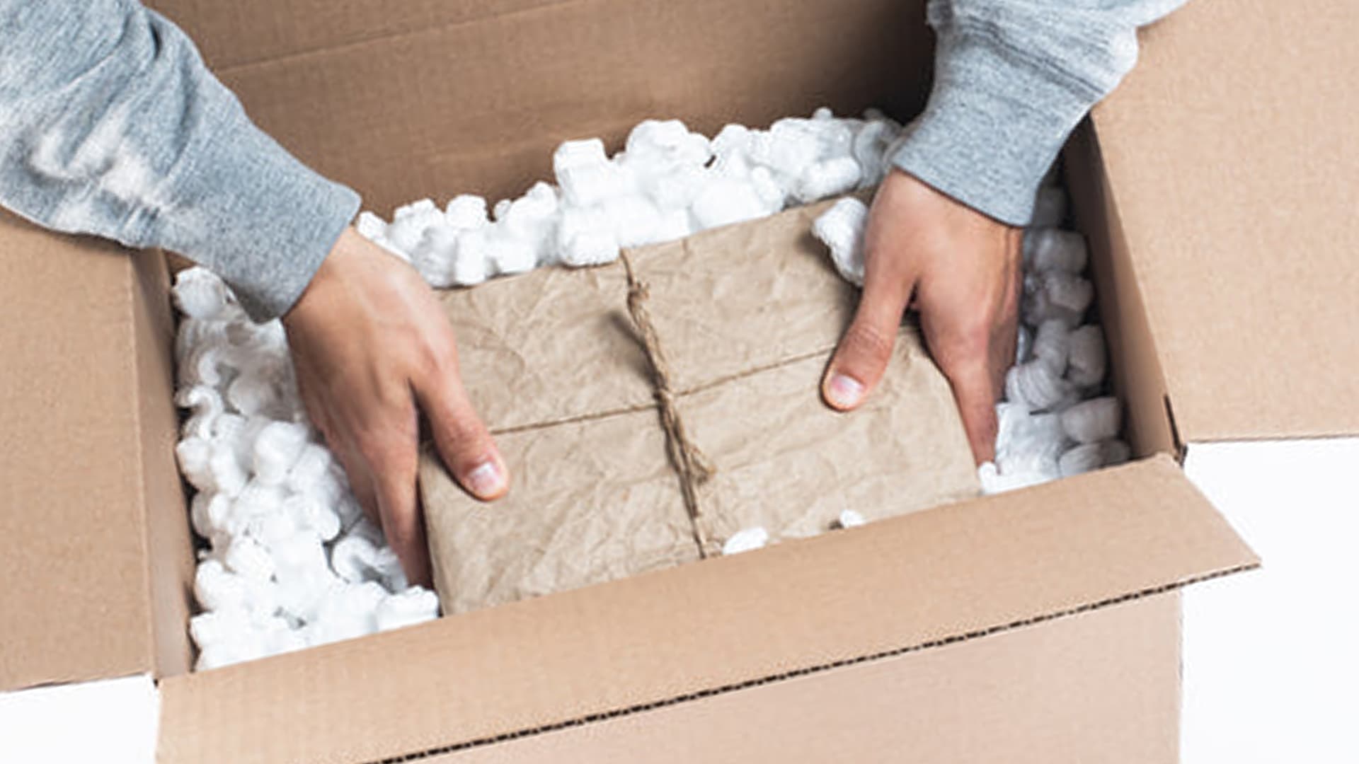 Hands in gray sleeves carefully pack wrapped items cushioned with white packing peanuts inside a brown cardboard shipping box, demonstrating protective packaging for shipping fragile goods.