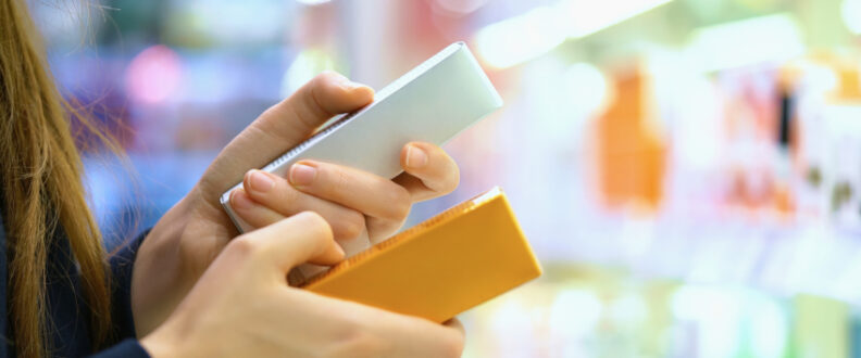 A person's hands holding two smartphones - one white and one orange/yellow - against a blurred colorful background, suggesting comparison shopping or mobile device selection.