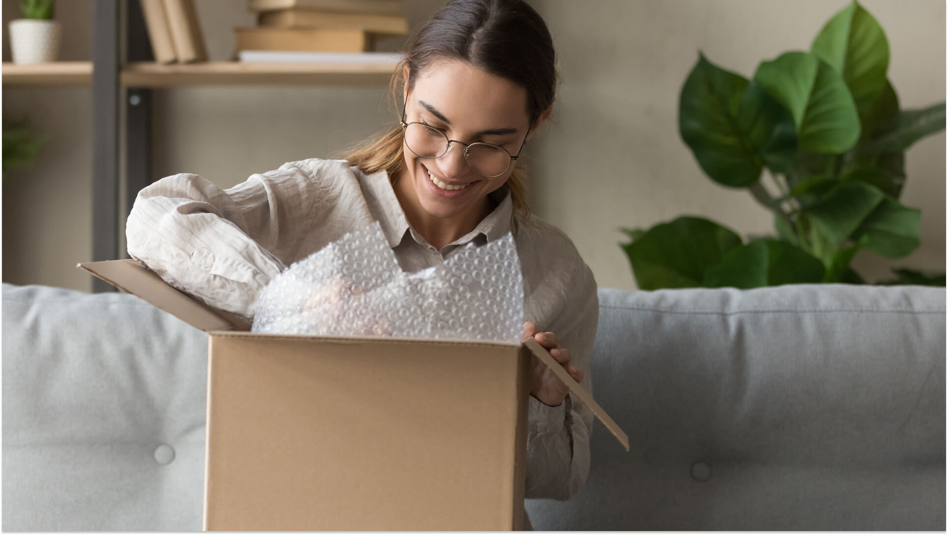 Happy woman sitting on couch opening cardboard e-commerce package with bubble wrap protective packaging in modern home living room
