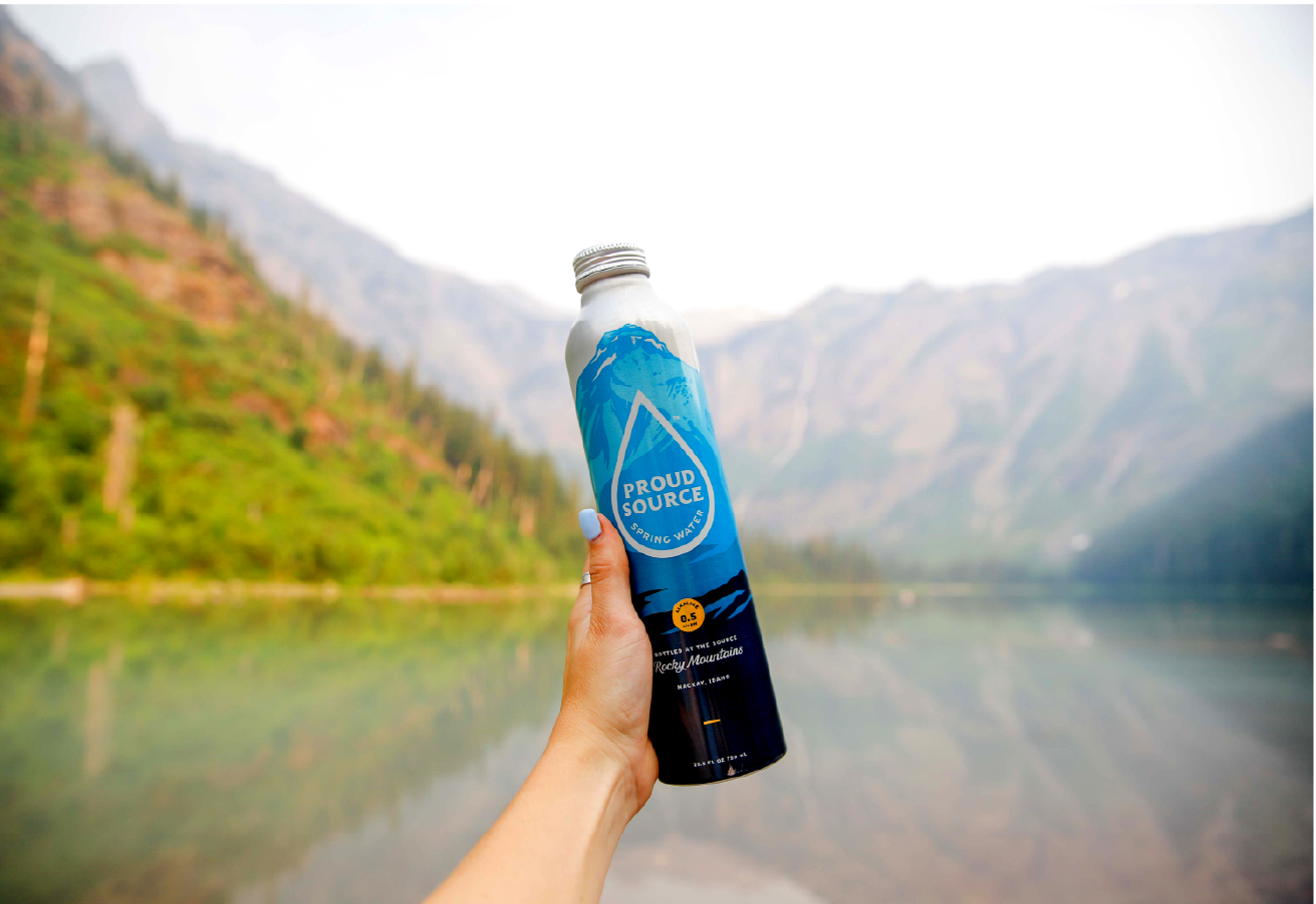 Hand holding Proud Source water bottle with mountain lake backdrop