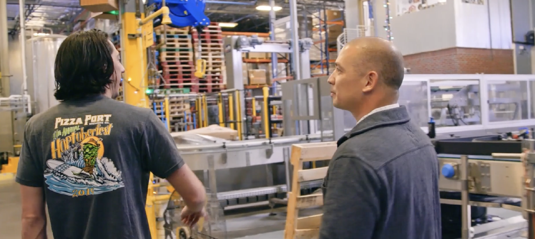 Two professionals touring manufacturing facility, observing automated packaging equipment and production line operations in industrial warehouse setting.