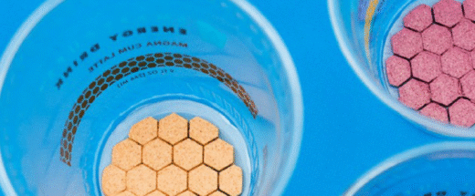 Overhead view of blue plastic cups containing hexagon-shaped crackers or supplements in different colors - orange, pink, and other varieties - arranged on a bright blue background