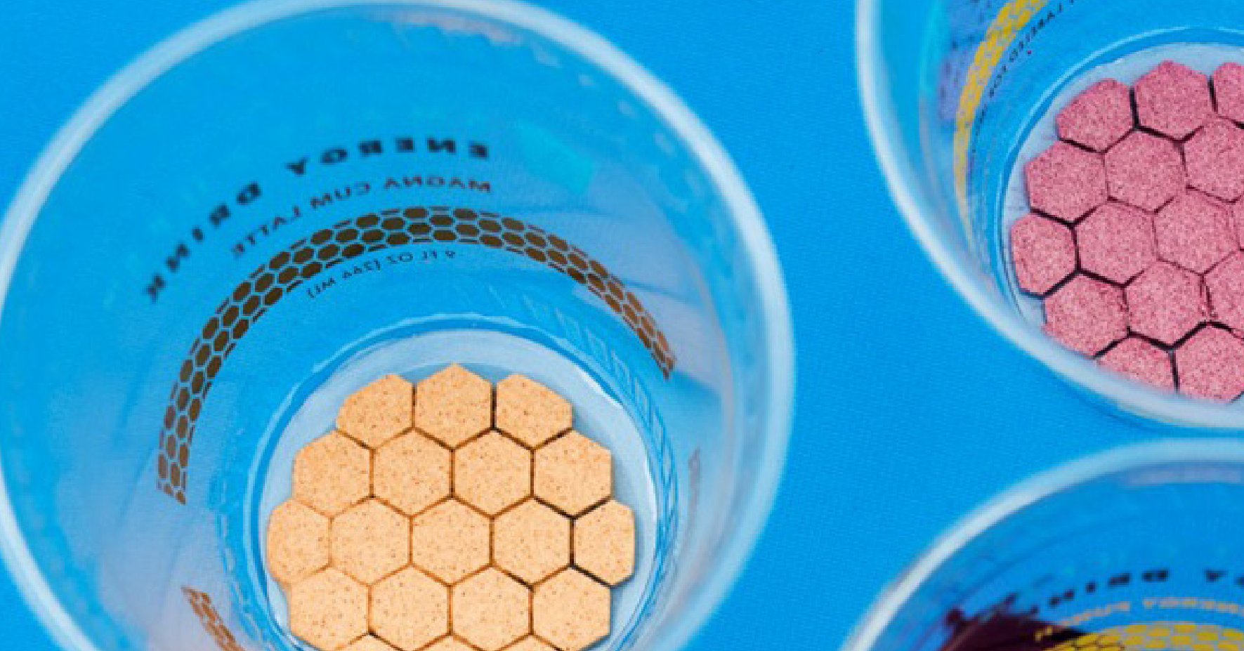 Overhead view of blue plastic cups containing hexagon-shaped crackers or supplements in different colors - orange, pink, and other varieties - arranged on a bright blue background