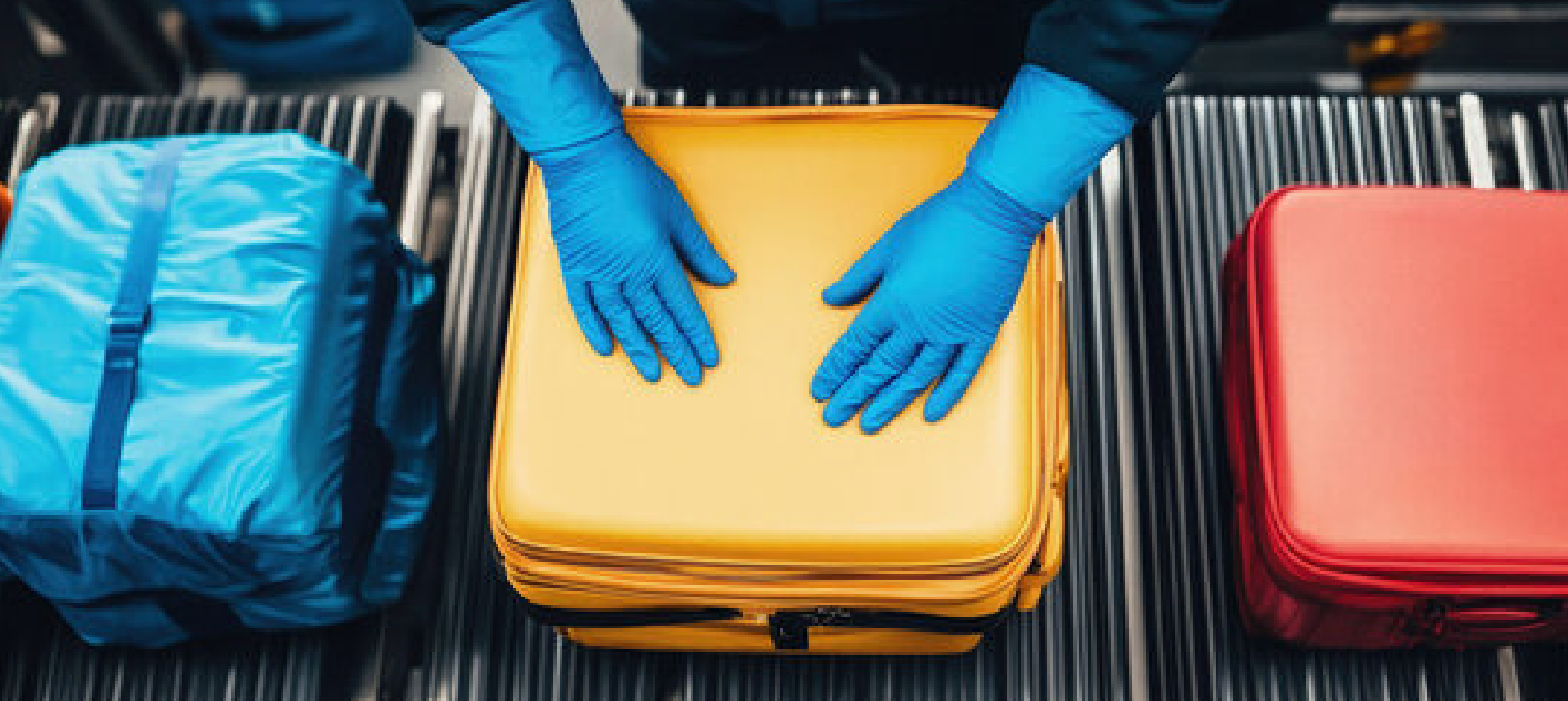 Aerial view of colorful luggage on a conveyor belt with gloved hands reaching toward a yellow suitcase, flanked by blue and red luggage cases
