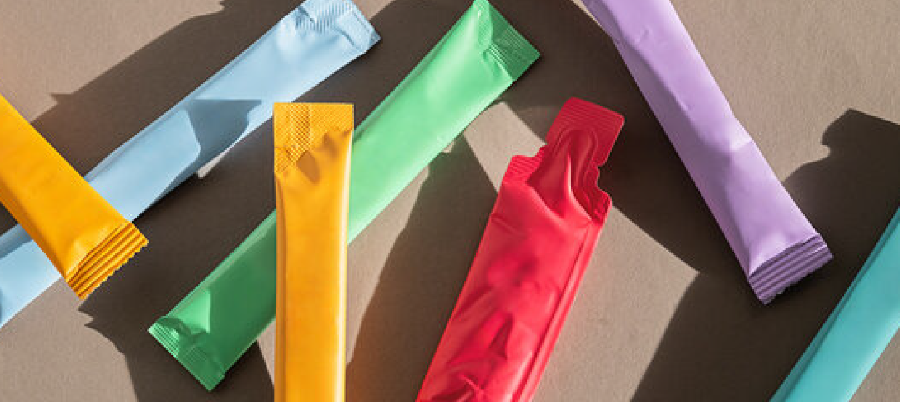 Colorful single-serve supplement or powder packets arranged on a dark surface, featuring stick packs in yellow, green, red, blue, and purple with textured honeycomb patterns