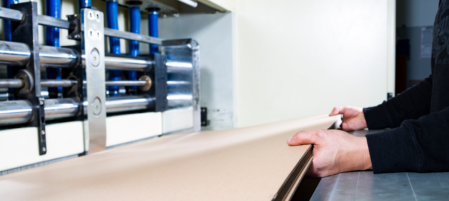 Worker's hands feeding a corrugated cardboard sheet through an industrial manufacturing roller machine.