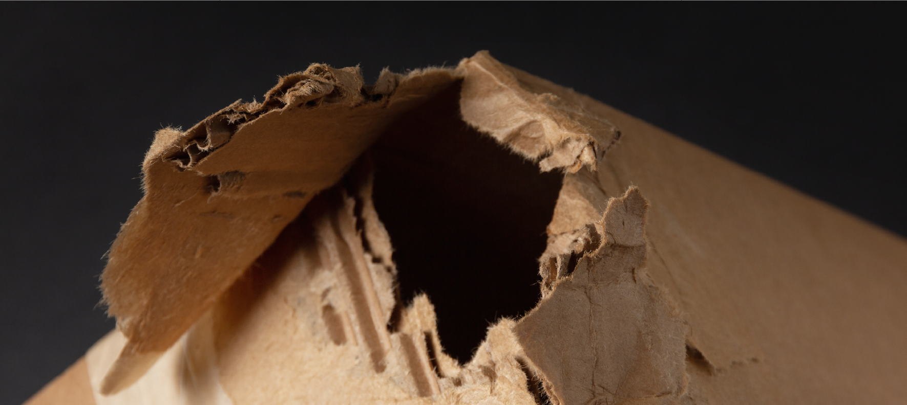 Damaged corrugated cardboard box corner with a large torn hole exposing the inner fluted layers against dark background.