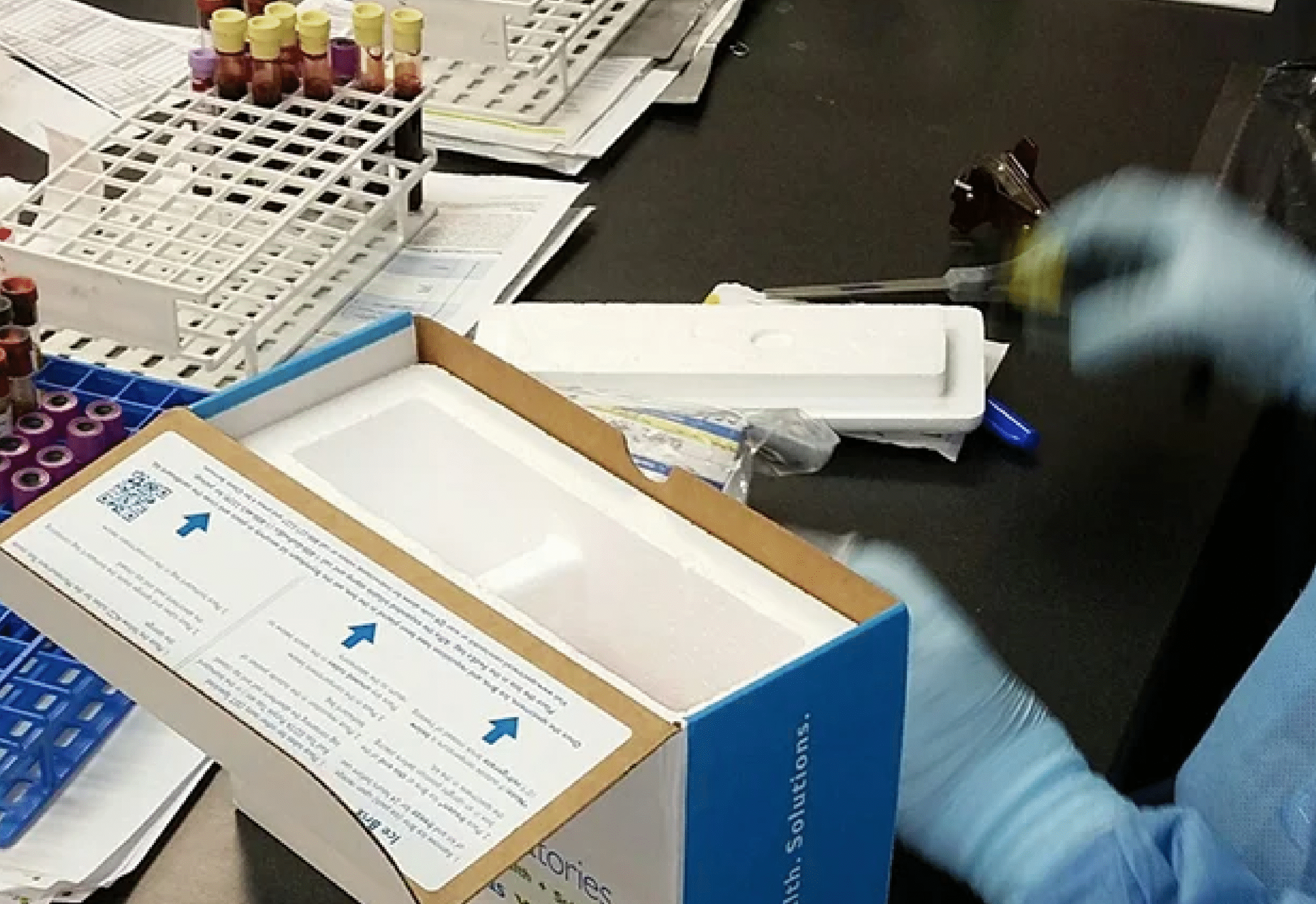 Laboratory technician in blue gloves handling medical specimen packaging with test tube racks nearby.