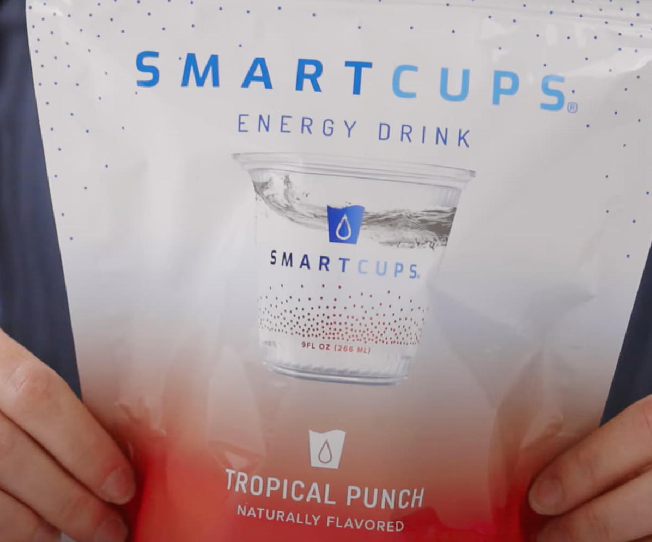 A person holding a white and red stand-up pouch of SmartCups Energy Drink in Tropical Punch flavor, naturally flavored, featuring a branded cup graphic on the packaging.