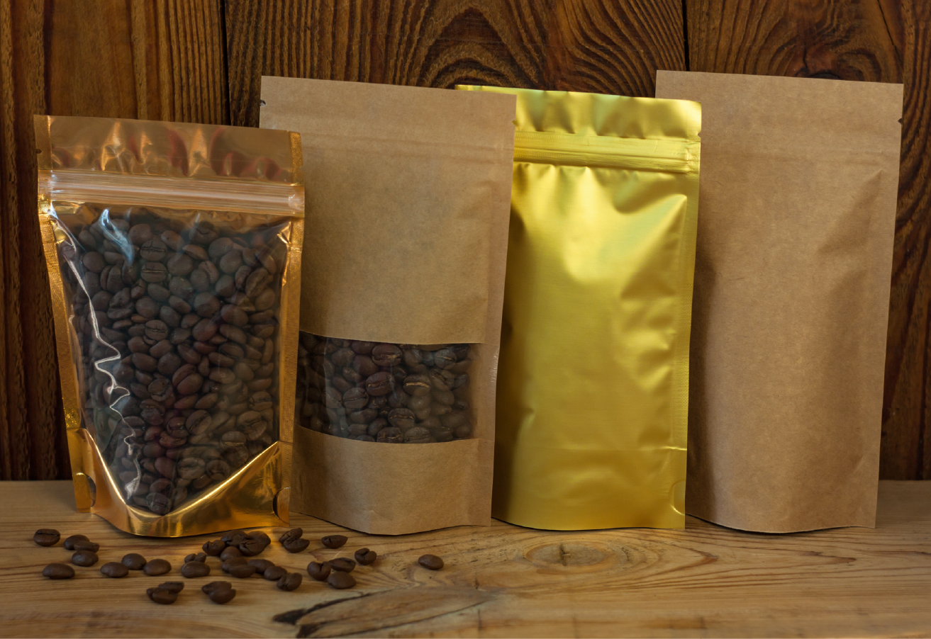 Four stand-up pouches in varying styles — clear/gold foil, kraft with window, solid gold foil, and plain kraft — displayed on a rustic wooden surface with scattered coffee beans in the foreground.