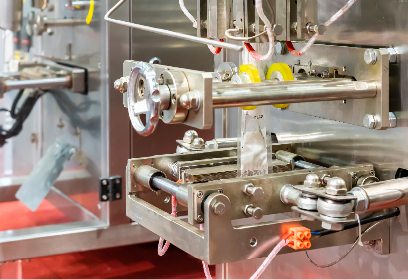 A close-up of industrial stainless steel pouch-filling and sealing machinery in operation, with mechanical arms, rollers, and a partially formed clear pouch visible.