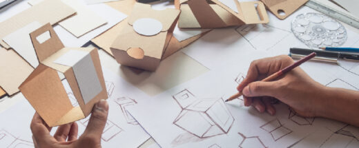 A designer's hands sketch box prototypes on paper beside handcrafted cardboard packaging models and drafting tools on a worktable.