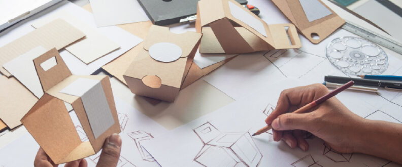 A designer's hands sketch box prototypes on paper beside handcrafted cardboard packaging models and drafting tools on a worktable.