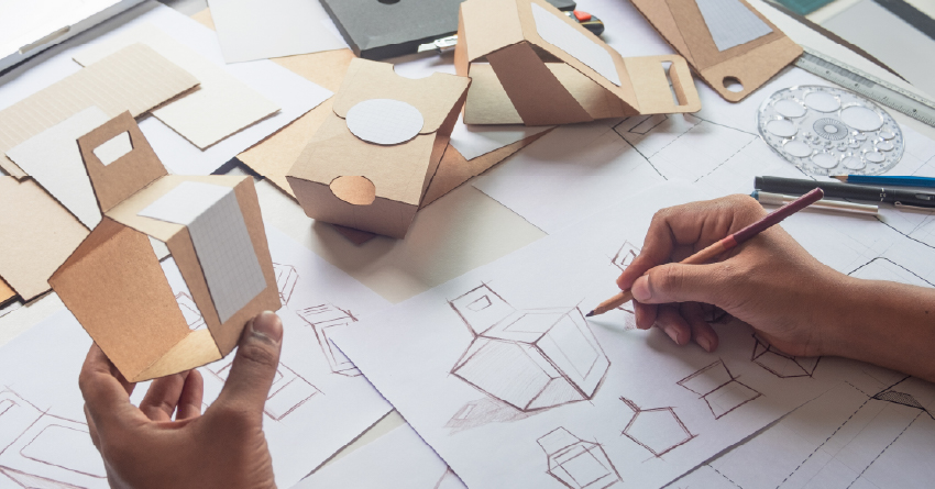 A designer's hands sketch box prototypes on paper beside handcrafted cardboard packaging models and drafting tools on a worktable.