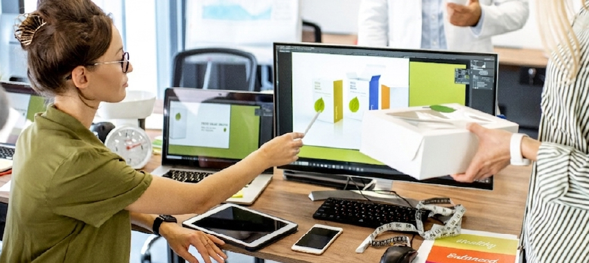 A female designer points to a 3D product packaging mockup on a monitor while a colleague holds a physical white box.