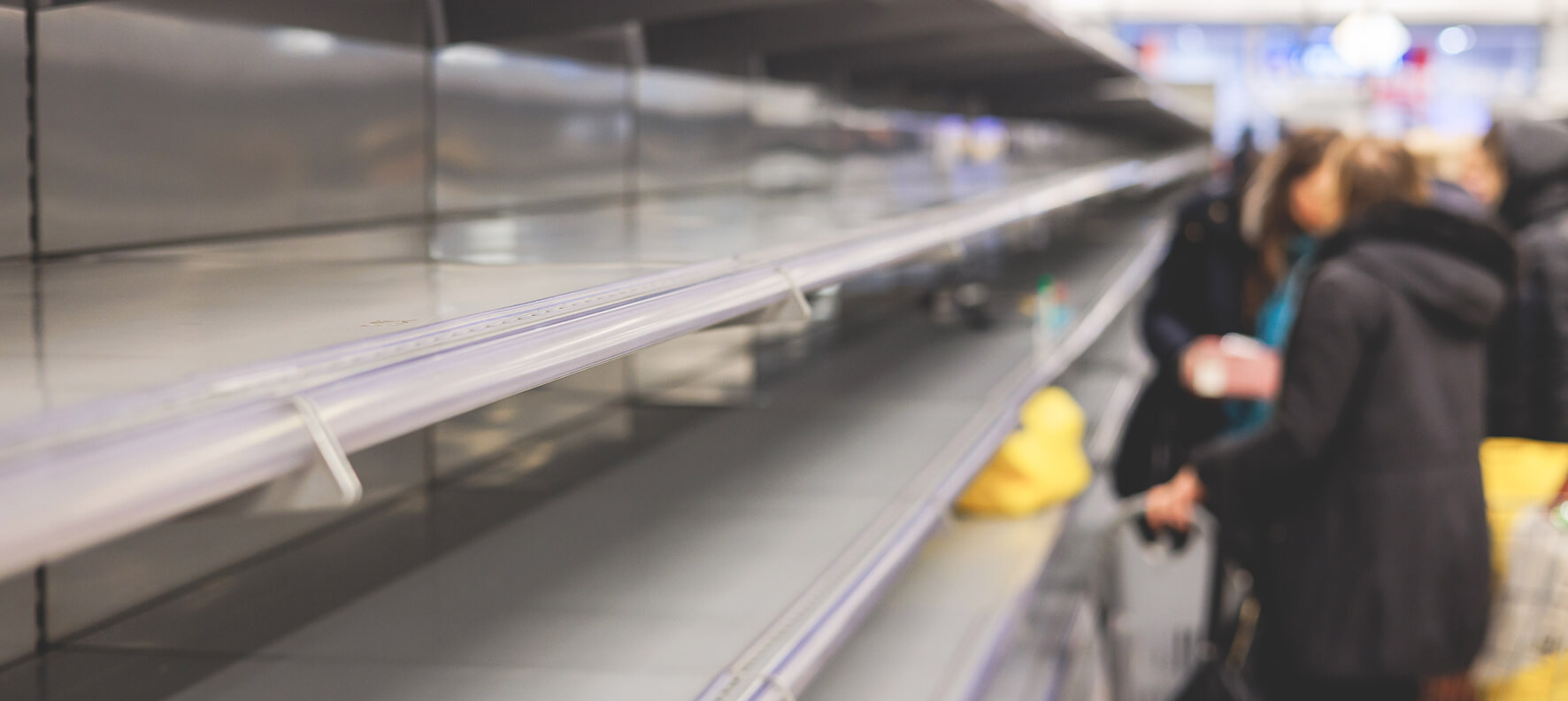 A shopper in a dark coat looks at nearly empty refrigerated shelves in a grocery store, illustrating a supply shortage.