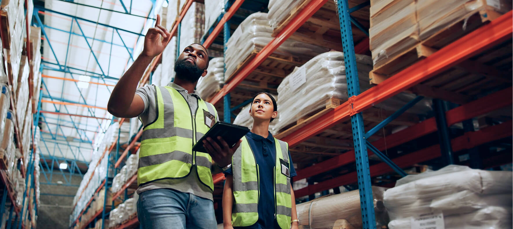 Two warehouse workers in bright yellow safety vests review inventory on a tablet while inspecting tall blue and orange shelving racks stocked with bagged goods.