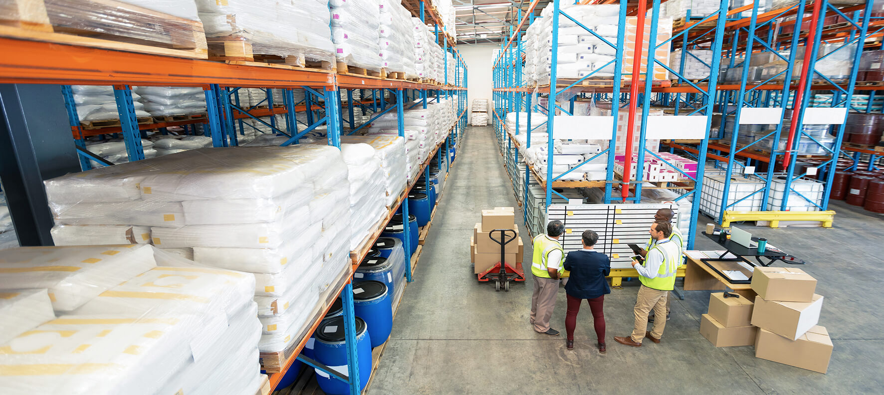 Three workers and a manager huddle around a tablet near a loaded pallet jack in a large warehouse stocked floor-to-ceiling with bagged materials and barrels.