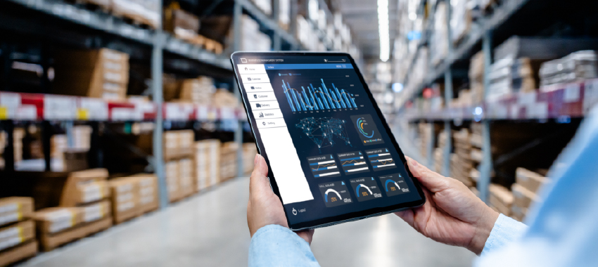 A worker in a blue shirt holds a tablet displaying a warehouse management dashboard with charts and data analytics.