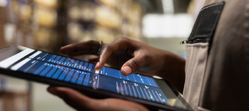 A close-up of a warehouse worker's hands tapping a tablet screen displaying blue inventory tracking charts and data.
