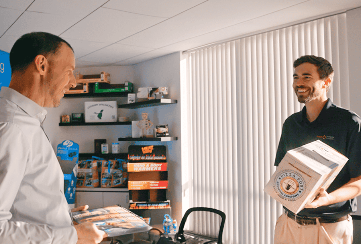 Two smiling men exchange branded packaging samples including a Pizza Port box in a bright office showroom displaying custom packaging products.
