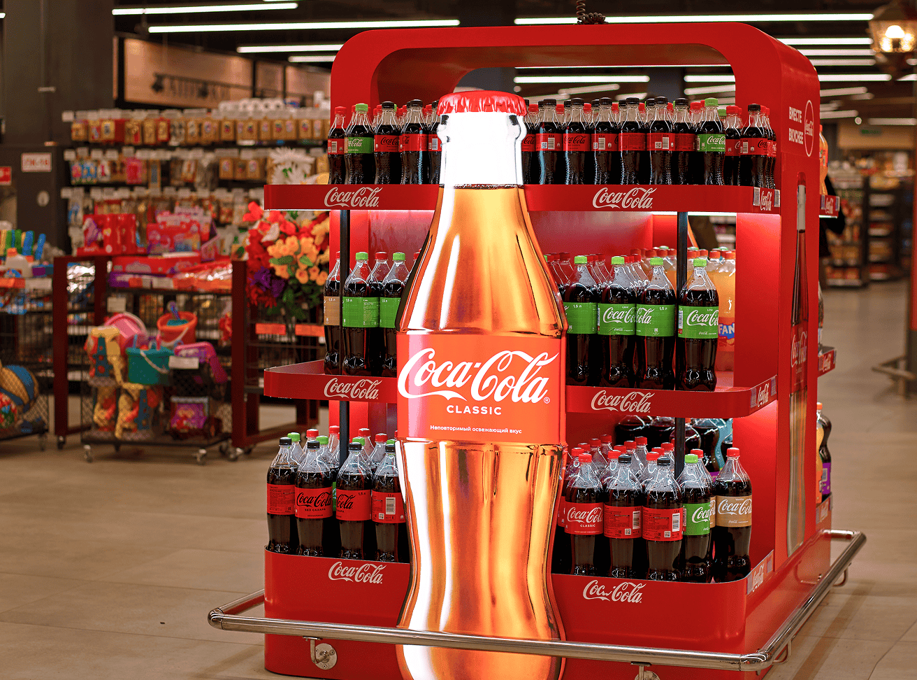 A bold red Coca-Cola Classic in-store display unit stocked with bottles and featuring a large gold decorative bottle centerpiece.