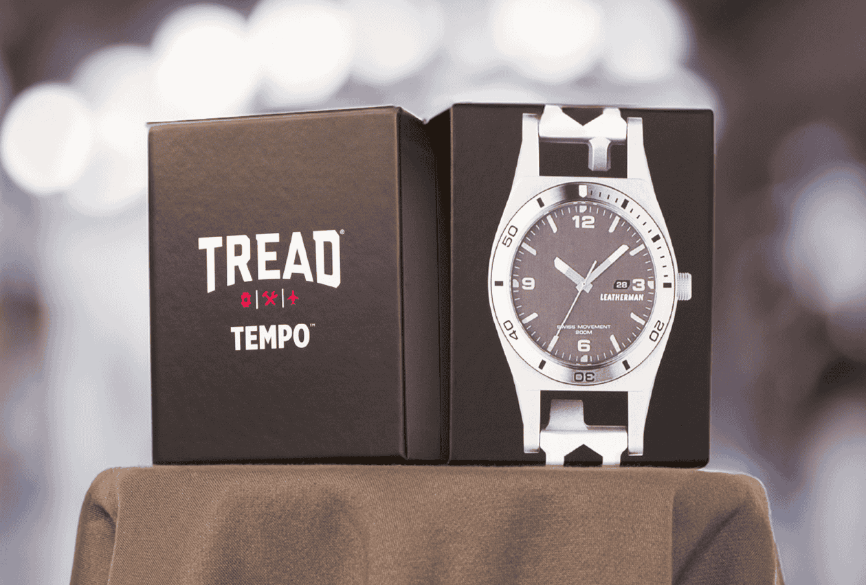 A Leatherman Tread Tempo watch displayed inside an open matte black branded gift box on a draped surface.