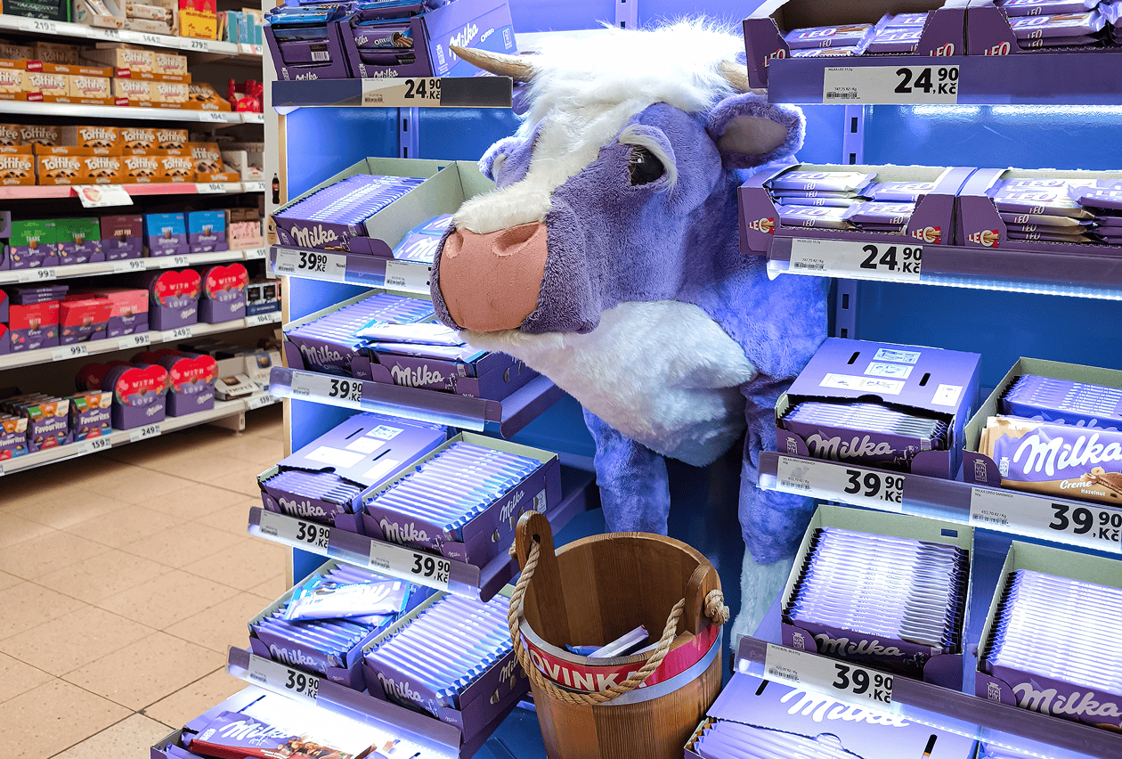 A purple plush cow mascot leans into a illuminated Milka chocolate display shelf stocked with candy bars in a retail store.