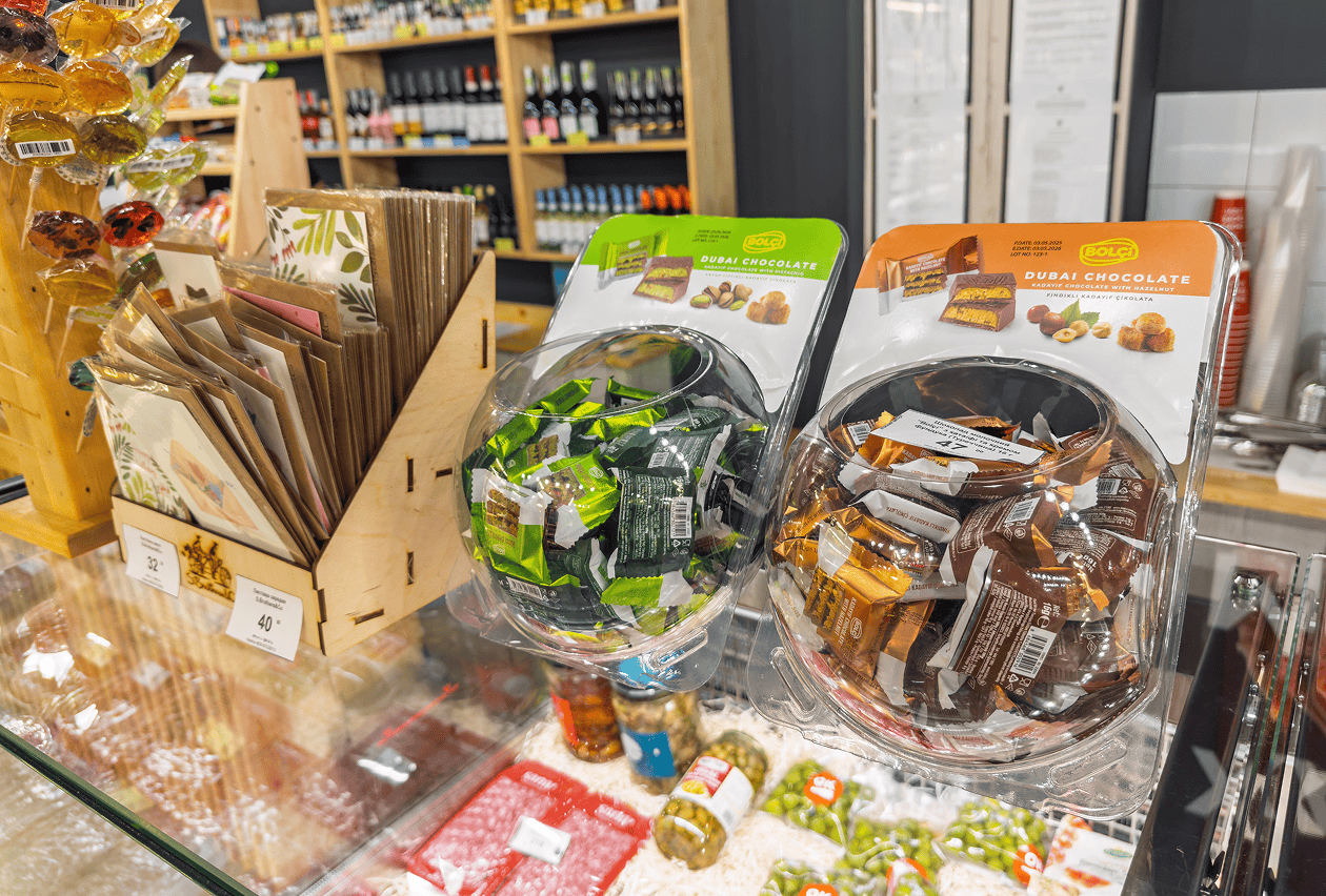 Two clear globe-shaped display bowls filled with wrapped Dubai Chocolate candies sit on a retail counter beside greeting cards and specialty food items.