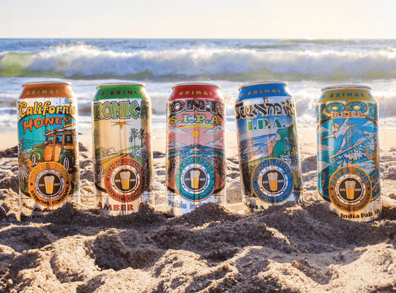 Five colorful Pizza Port Brewing Company beer cans with surf-themed artwork stand upright in beach sand with ocean waves behind them.