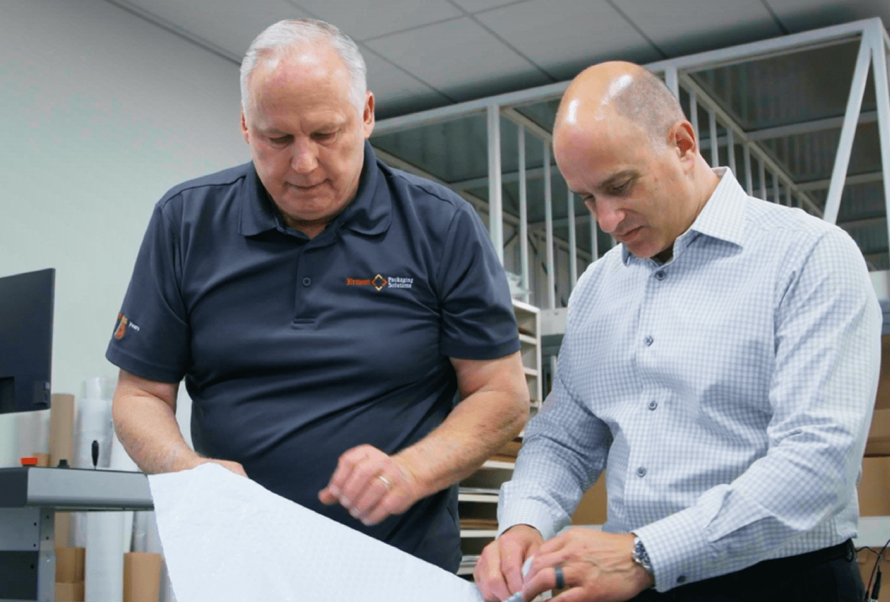 Two men in a packaging facility lean over a worktable to review a large white sheet of packaging material together.