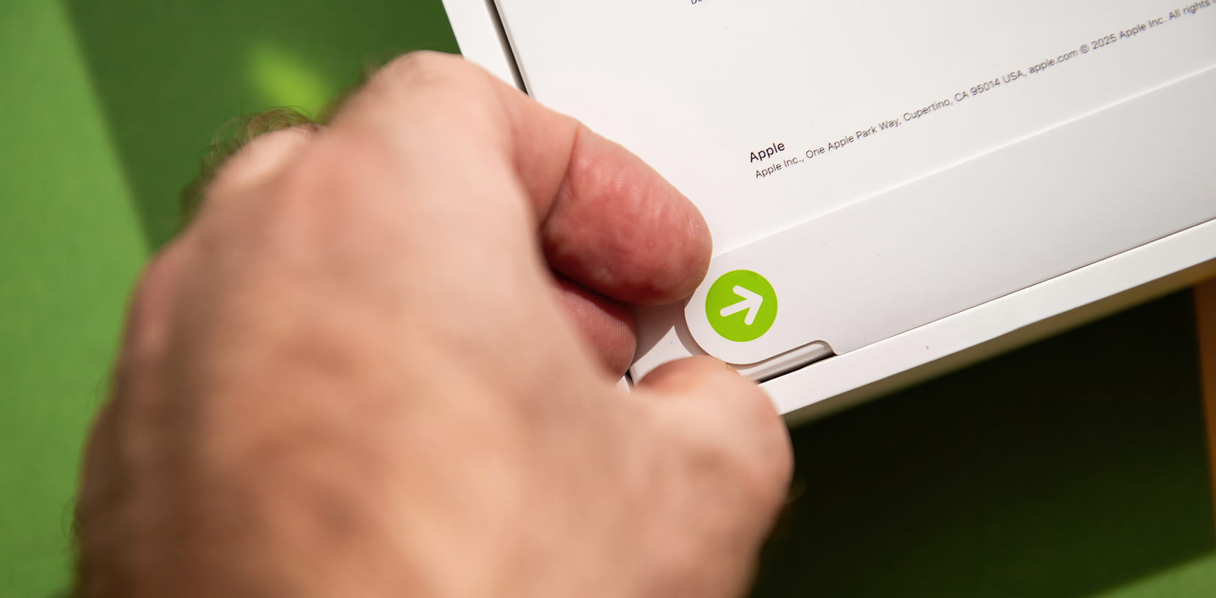 A hand opens an Apple product box, revealing a green arrow sticker on white packaging.