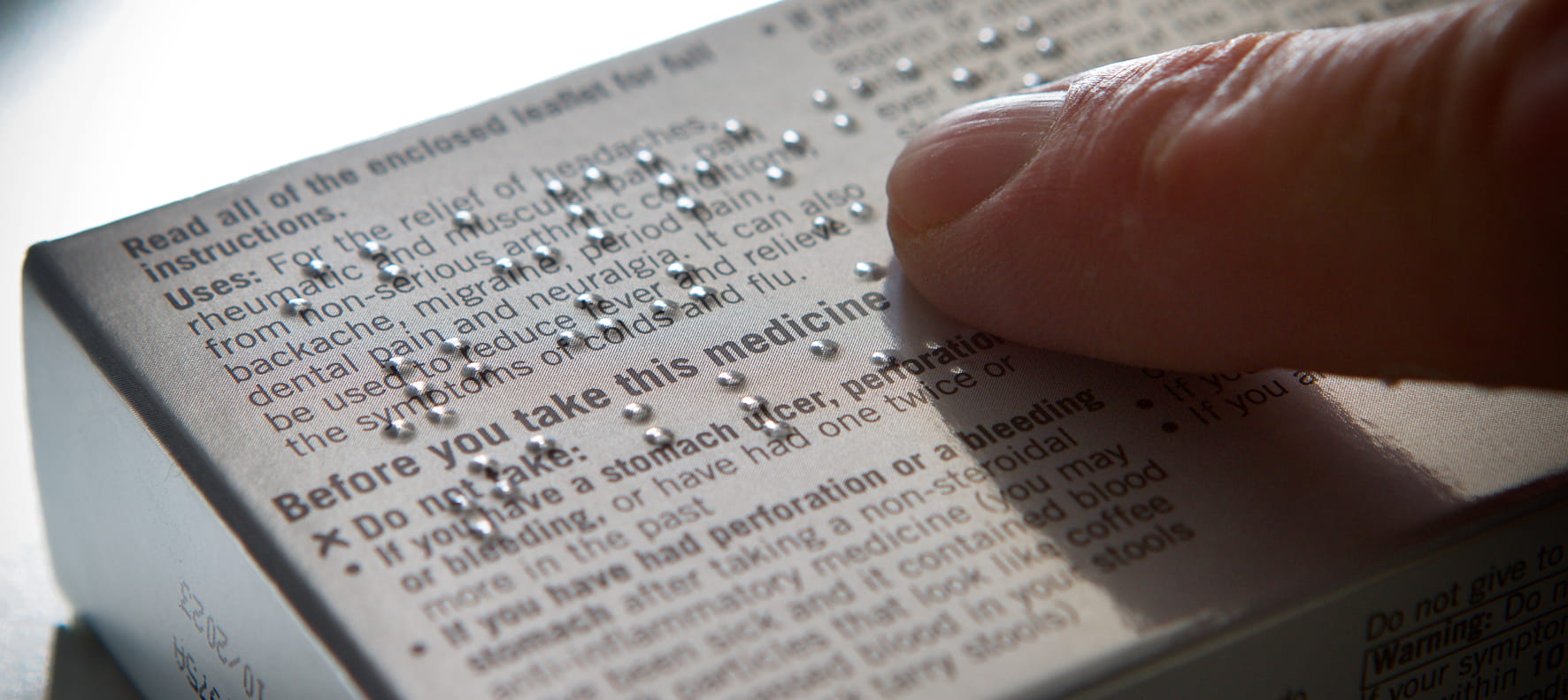 A finger reads Braille dots on a medicine box displaying drug uses and warnings.