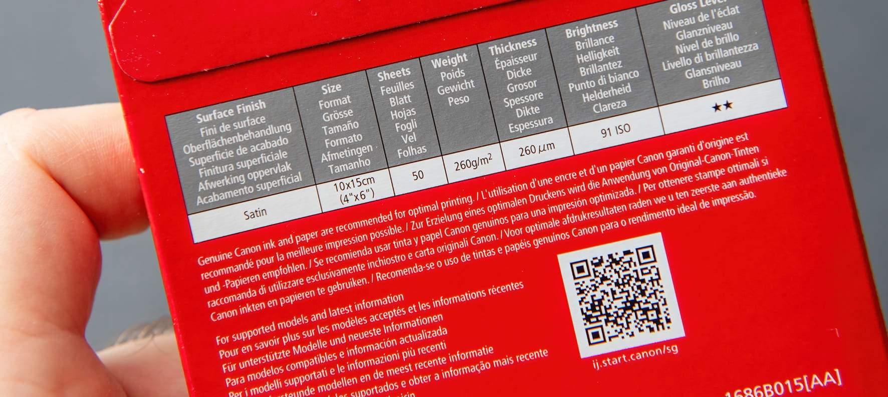 A hand holds a red Canon photo paper box showing multilingual specs and a QR code.