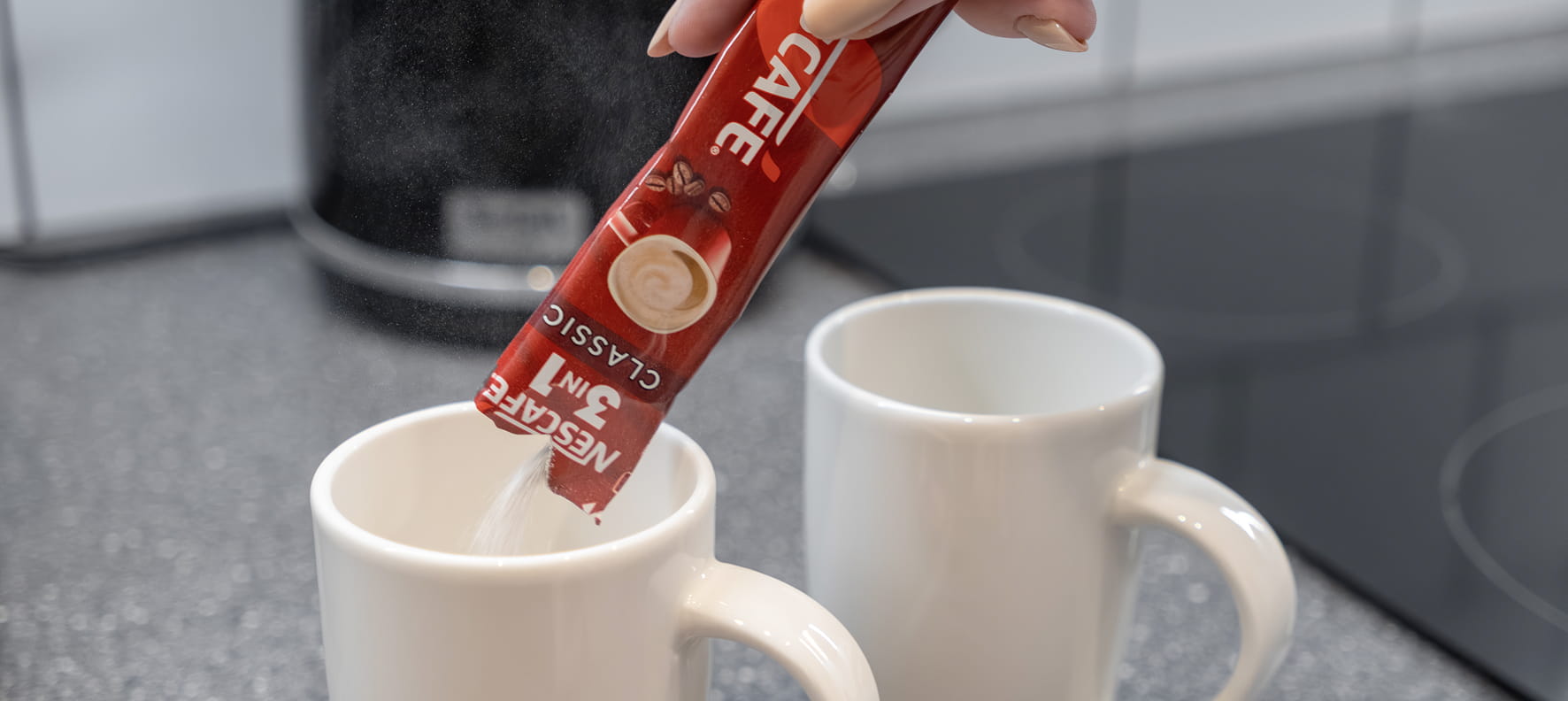 A hand pours a Nescafé 3-in-1 Classic stick packet into a white mug.