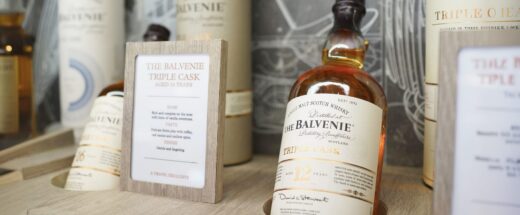 The Balvenie Triple Cask 12 Year single malt scotch whisky bottle displayed on a wooden retail shelf alongside cylindrical tube packaging and a branded tasting note card.