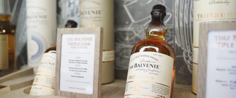 The Balvenie Triple Cask 12 Year single malt scotch whisky bottle displayed on a wooden retail shelf alongside cylindrical tube packaging and a branded tasting note card.