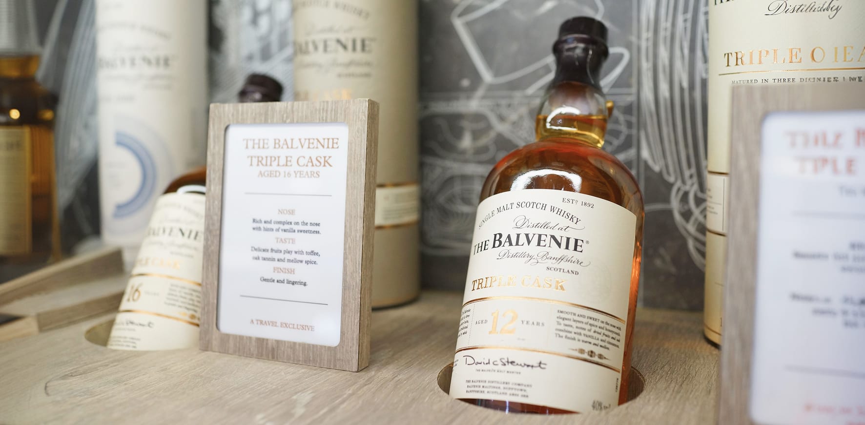 The Balvenie Triple Cask 12 Year single malt scotch whisky bottle displayed on a wooden retail shelf alongside cylindrical tube packaging and a branded tasting note card.