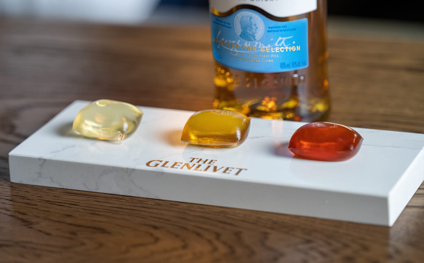 Three whisky-filled edible capsules in yellow, amber, and red displayed on a branded marble tasting board beside a bottle of The Glenlivet American Oak Selection.