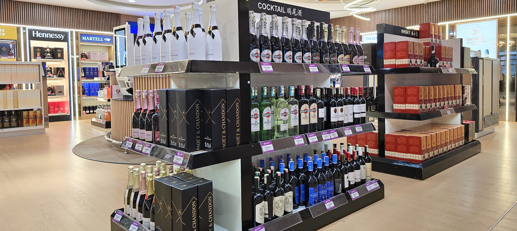 A duty-free retail display featuring Moët & Chandon, Martini, and whisky brands arranged across tiered shelving units in an airport store setting.