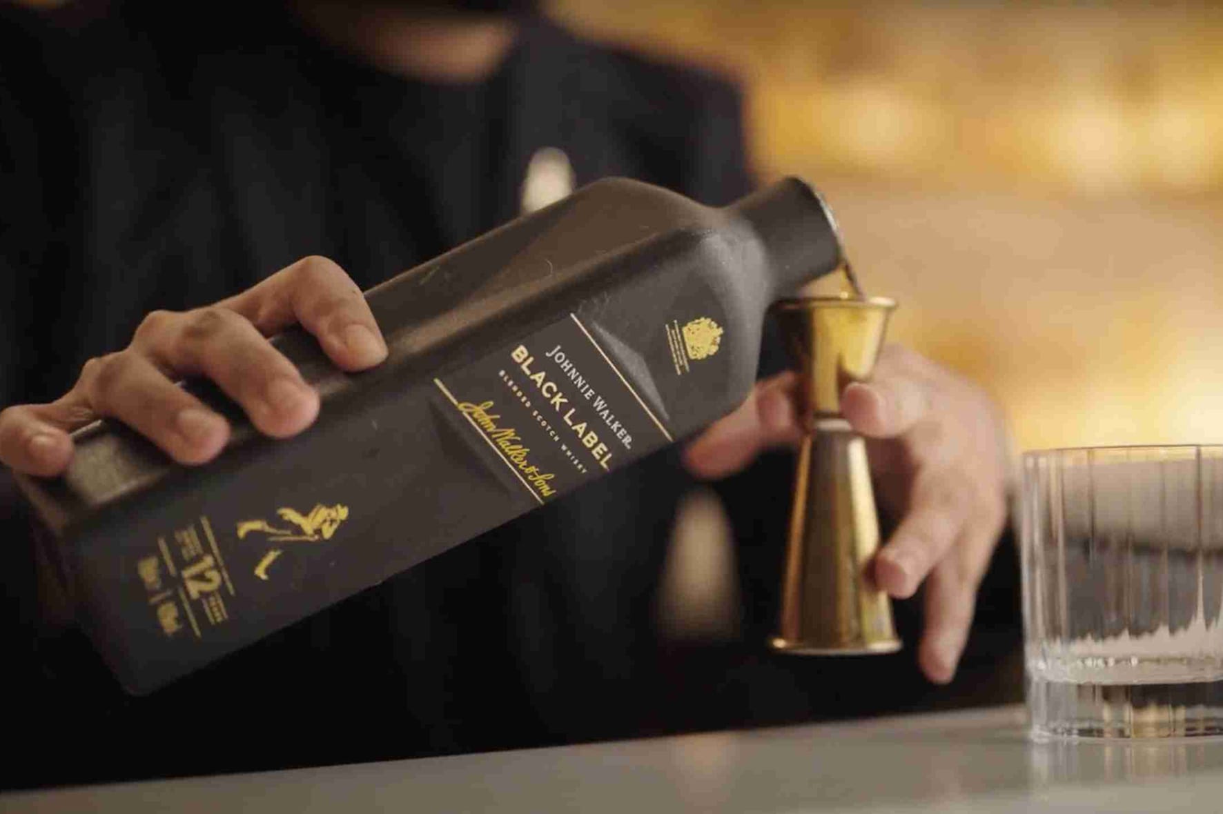 A bartender pouring Johnnie Walker Black Label blended scotch whisky into a brass jigger at a dimly lit bar.
