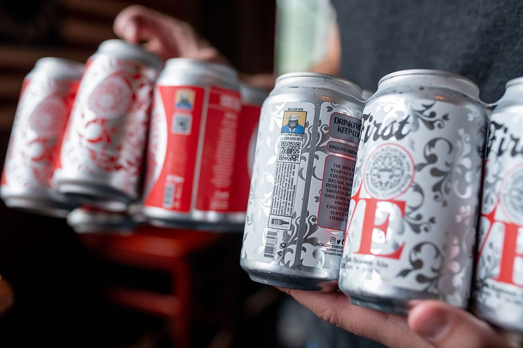 Two hands holding multiple craft beer cans including limited edition designs from McAllister Brewing, featuring QR codes and ornate label artwork.