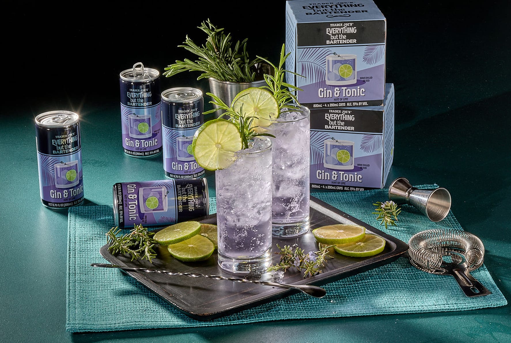 Trader Joe's Everything But the Bartender canned gin and tonic cocktails displayed alongside their multi-pack box packaging, garnished with lime and rosemary for styling.