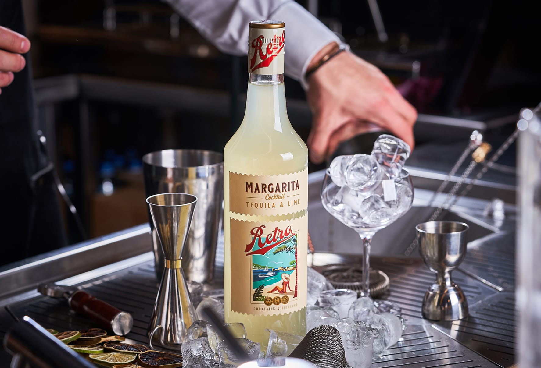 A bottle of Sodiko’s Retro line margarita cocktail mix surrounded by bar tools including a jigger, shaker, and coupe glass filled with ice.
