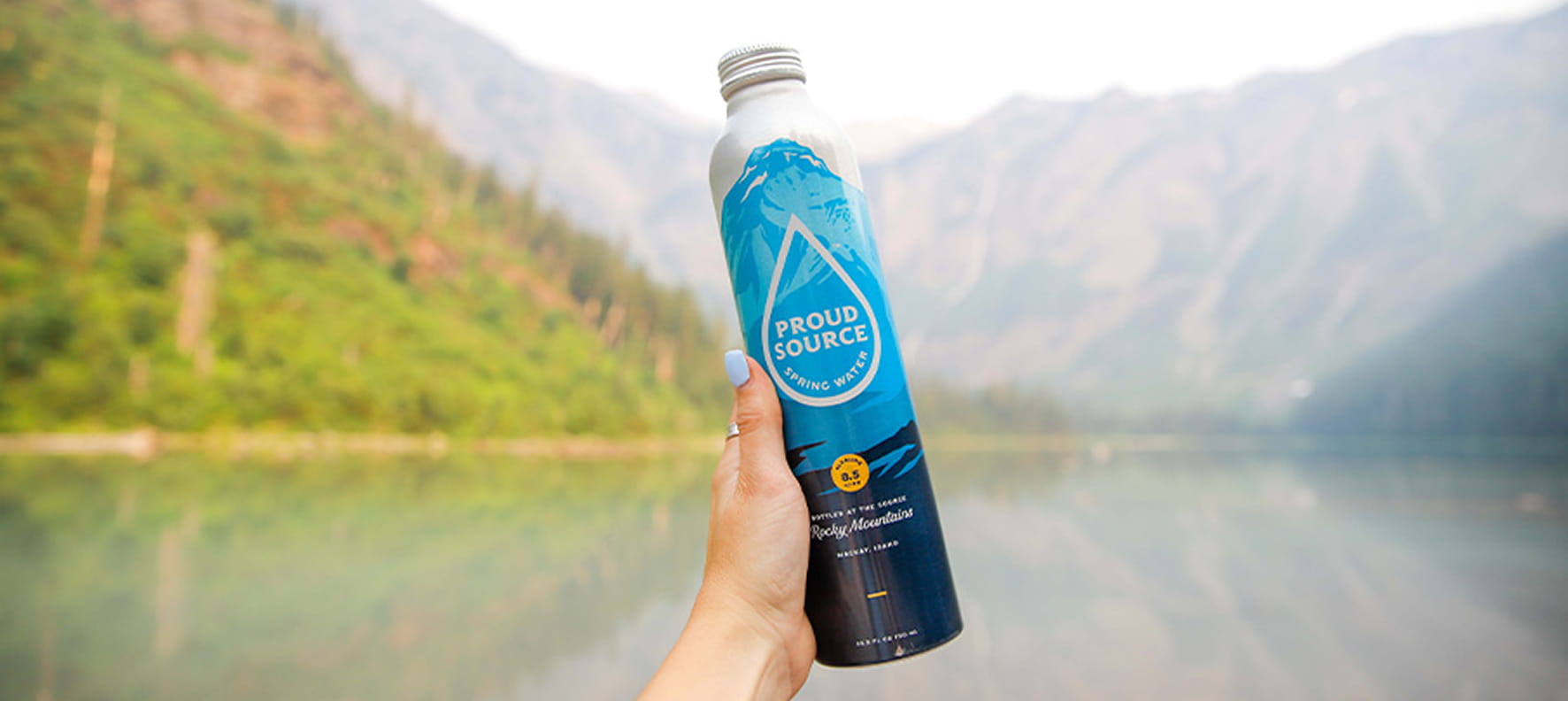 A hand holds a Proud Source Rocky Mountains aluminum spring water bottle against a misty mountain lake backdrop.