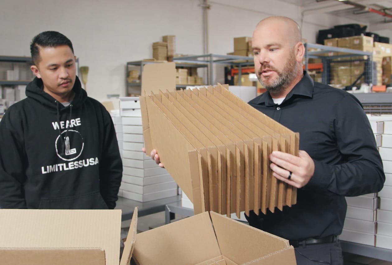 Two warehouse workers inspect a stack of flat-packed corrugated cardboard box dividers.