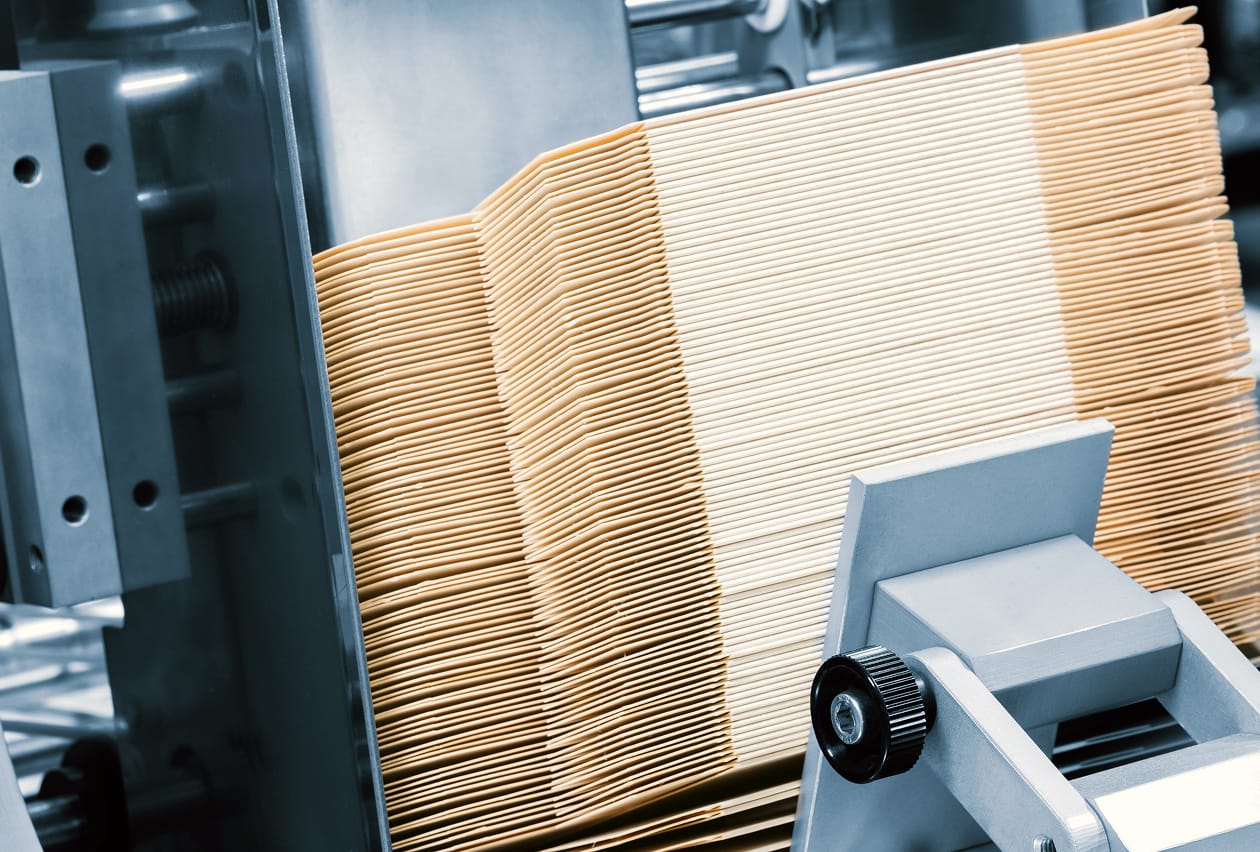 Stacks of flat kraft cardboard sheets feeding through an industrial die-cutting machine.